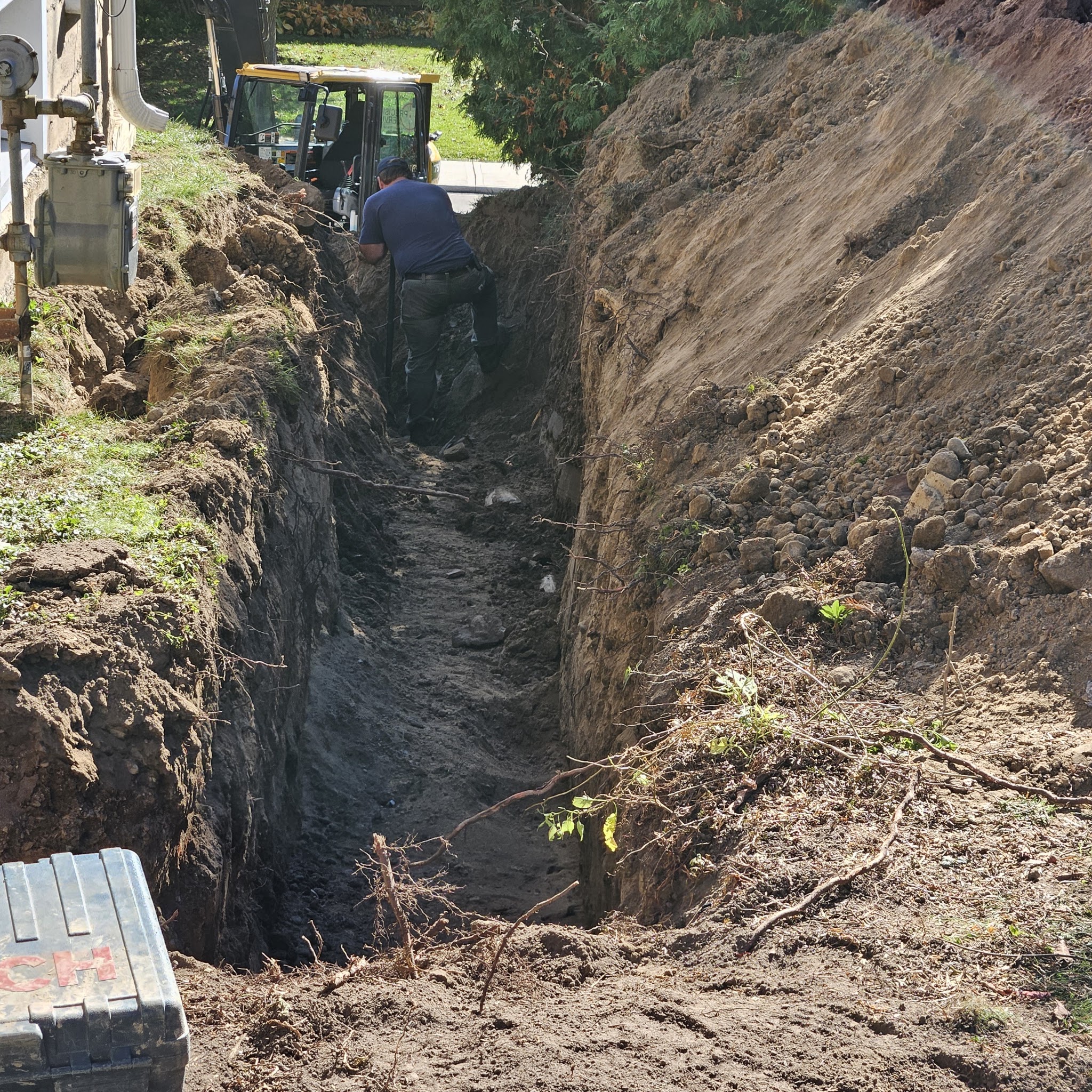 A crew member working in a deep excavated trench alongside a mini excavator during a sewer line replacement